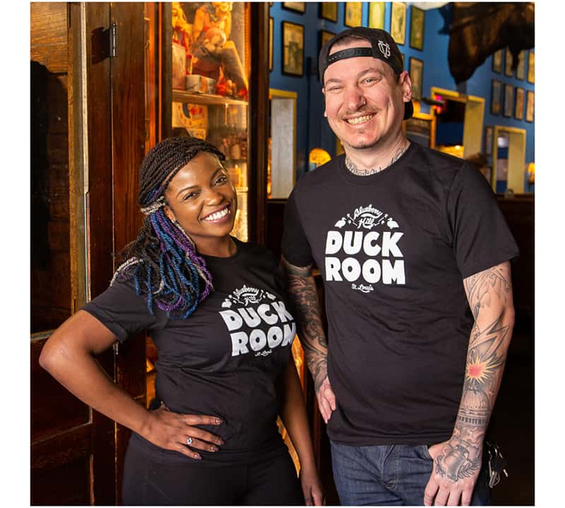 A black woman and a white man wear black t shirts featuring the Duck Room live music venue logo. The logo is made up of hand-drawn fat letters saying "Duck Room" in white on a black background. A script logo of Blueberry Hill is on top of it and St. Louis in script is below. There are two ducks on either side.