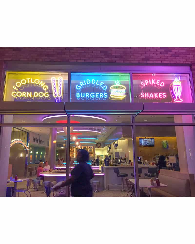 Three neon signs in the window of a diner featuring a yellow sign that says Footlong Corn Dogs and has corn dogs in it, a blue one that says Griddled Burgers with a yellow, green, and red hamburger next to it, and a pink one that says Spiked Shakes with a pink and yellow milkshake next to it.