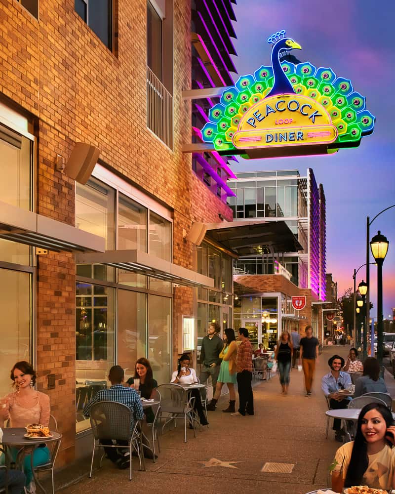 An outdoor sidewalk scene showing people dining on the patio in front of Peacock Diner. Above them is the huge 11-ft neon sign of a peacock with blue, green, and yellow feathers fanning out behind the slender curved neck. Peacock Loop Diner is in retro neon letters on the sign.