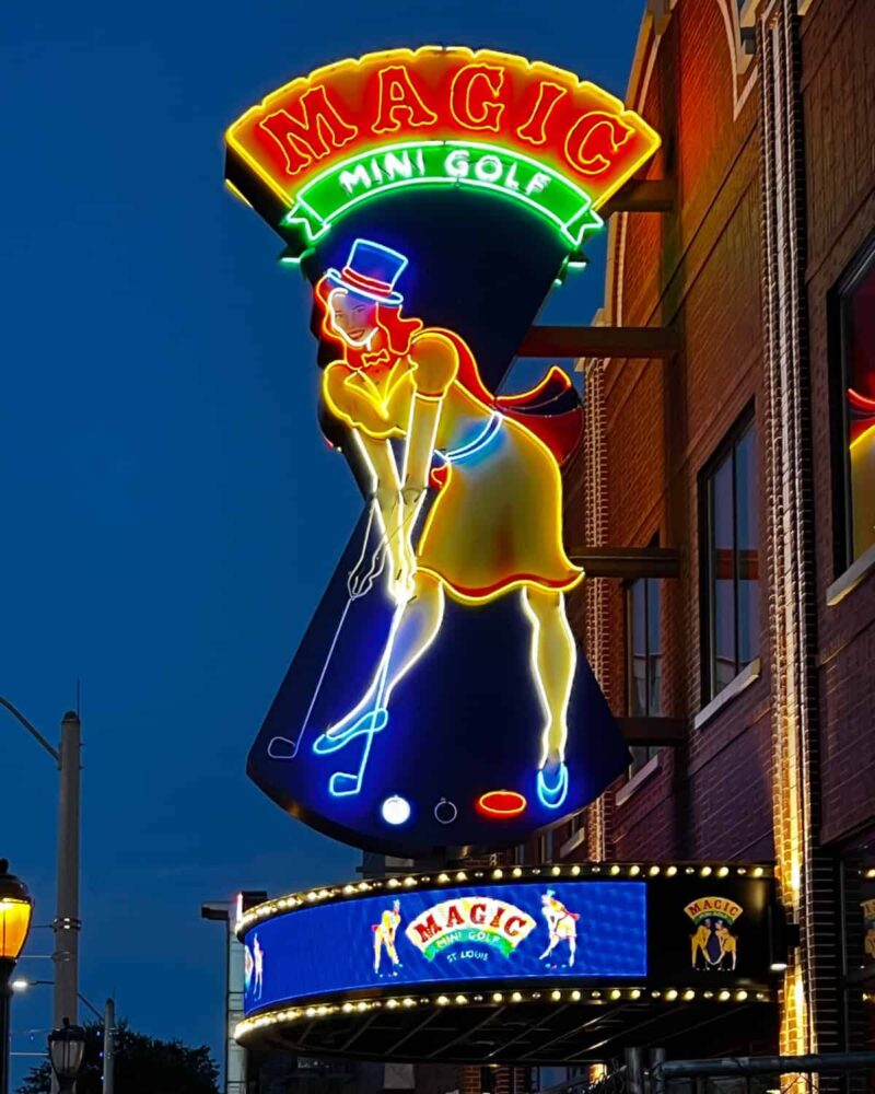 Huge 21' Magic Mini Golf neon sign at dusk. Big red neon circus style letters on a curved yellow neon-outlined background with a woman in yellow neon putting mini golf. She is wearing a yellow dress, a light blue magician's hat, and a red cape.
