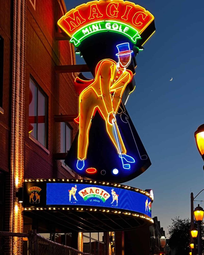 Huge 21' Magic Mini Golf neon sign at dusk. Big red neon circus style letters on a curved yellow neon-outlined background with a man putting mini golf. He is wearing a yellow tuxedo, a light blue magician's hat, and a red cape, all of which are outlined in neon.